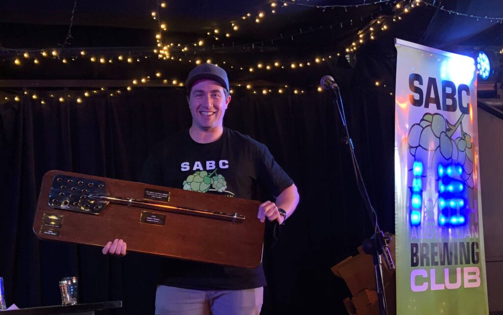 Brendan in SABC t-shirt holds the SABSISA trophy, a silver mash paddle mounted on a wooden board